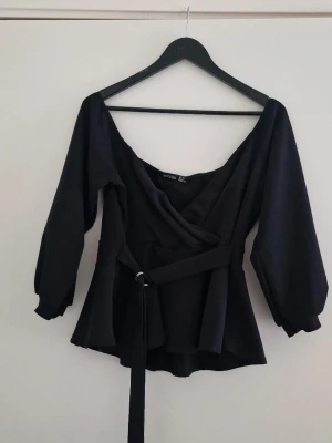 Svart omlottblus med bälte från Boohoo - Stilren svart off shoulder topp från Boohoo med långa puffärmar och markerad midja tack vare det medföljande bältet. Blusen har djup v-ringning och är tillverkad i ett mjukt, vävt material som ger en snygg siluett. Som ny. Storlek 44/46, passar 44. Elastisk. 