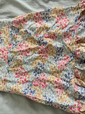 Blommig kjol från Gina Tricot - Söt blommig kjol från Gina Tricot i vitt med små blommor i rosa, blått och gul. Kjolen har volangdetaljer och är tillverkad i mjuk bomull, perfekt för en fräsch och somrig look. Säljer då den är för liten