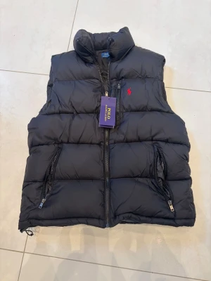 Svart dunväst från Polo Ralph Lauren - Snygg svart dunväst från Polo Ralph Lauren med dragkedja framtill, två fickor med dragkedja och klassisk röd logga på bröstet. Västen har hög krage och quiltad design som ger en sportig och stilren look. Perfekt för lager-på-lager och streetstyle. Pris kan diskuteras 