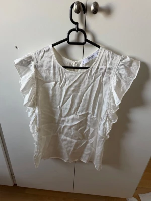 Vit volangblus från Mango - Snygg vit blus från Mango med volangdetaljer längs sidorna och korta ärmar. Blusen har rund halsringning och är tillverkad i ett lätt och luftigt material, perfekt för varmare dagar.