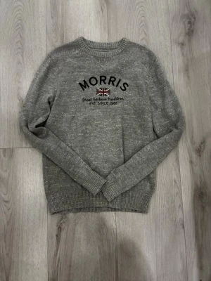Grå stickad tröja från Morris - Snygg grå stickad tröja från Morris med broderad text och brittisk flagga på bröstet. Tröjan har rund halsringning, ribbade muddar och är perfekt för en stilren och avslappnad look.