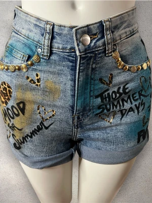 Unika blå denimshorts - Säljer ett par blå denimshorts med hög midja, dekorerade med guldfärgade nitar vid fickorna. Shortsen är kostumiserade och handmålade av designern DGR, vilket gör dem till en helt unik och exklusiv piece. De har leopardmönstrade, handmålade texter samt detaljer i svart och guld. Perfekta för dig som vill sticka ut i sommar!