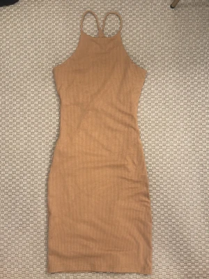 Beige ribbad midiklänning - Snygg beige ribbad klänning med smala axelband och hög hals. Klänningen har en kroppsnära passform och är ärmlös, perfekt för varma dagar eller att styla med en jacka. Materialet är stretchigt och mjukt för extra komfort.