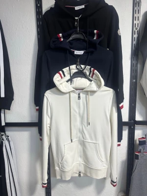 Svart vit blå moncler hoodie med dragkedja och ränder - Svart Vit blå  hoodie från Moncler med dragkedja framtill, huva med dragsko och röda samt marinblå ränder på ärmslut och huva. Två fickor framtill och ribbade muddar. Perfekt för en sportig och stilren look.