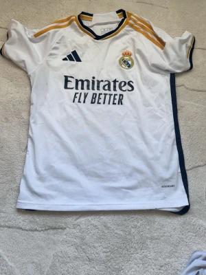 Real Madrid matchtröja vit Adidas - Vit Real Madrid matchtröja från Adidas med gula och mörkblå detaljer. Tröjan har klubbmärke på bröstet, Adidas-logga och sponsortryck 'Emirates Fly Better' framtill. Tillverkad i lätt och ventilerande material med Aeroready-teknologi.