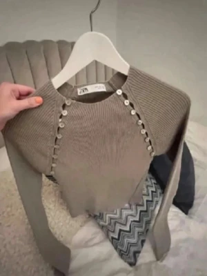 Beige stickad tröja med knappar Zara - Trendig beige stickad tröja från Zara med dekorativa knappar längs axlarna. Tröjan har rund halsringning och långa ärmar. Perfekt för lager-på-lager och passar till både jeans och kjol.