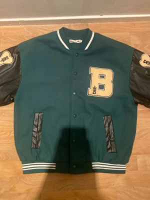 Ealey fushi  - Snygg varsity-inspirerad overshirt i mörkgrönt tyg med svarta ärmar i skinnimitation. Stor gul B-patch på bröstet och detaljer på ärmarna. Två svarta fickor framtill och ribbade muddar med vita ränder. Perfekt för en trendig streetwear-look.