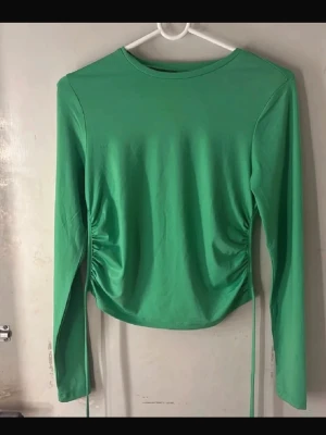 Långärmad topp - Trendig grön långärmad topp med snörning i sidorna och öppen rygg. Toppen har en tight passform och är tillverkad i ett mjukt, stretchigt material som känns skönt mot huden. Perfekt för dig som vill sticka ut med en cool detalj på ryggen. Perfekt till sommar!