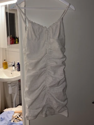 Vit kort klänning med broderade detaljer - Söt vit kort klänning med smala axelband och rynkade detaljer framtill och baktill. Klänningen har broderade hålmönster och en figurnära passform som ger en feminin look. Perfekt för varma dagar och somriga tillfällen.
