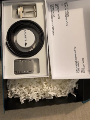 Svart bälte med silverdetaljer Lacoste - Säljer ett stilrent svart bälte från Lacoste med silverfärgat spänne och extra platta med Lacoste-logga. Bältet är i slätt material och kommer i originalbox med krokodiltryck. Perfekt för att lyfta din outfit med en klassisk accessoar.