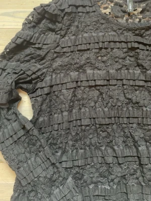 Svart spetsblus med volangdetaljer - Snygg svart blus från ONLY i spets med långa ärmar och volangdetaljer över hela toppen. Blusen har rund halsringning och är genombruten med ett feminint uttryck. Perfekt för dig som gillar en trendig och elegant look. 