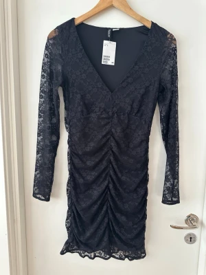 Svart spetsklänning med v-ringning - Svart kort klänning från H&M i spets med v-ringning och långa ärmar. Klänningen har rynkade detaljer framtill och är figurnära. Perfekt för dig som gillar en elegant och trendig look. 