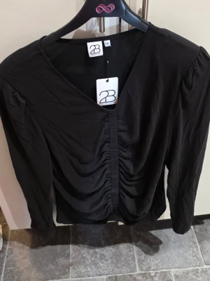 Svart blus med rynk framtill 2-Biz - Stilren svart blus från 2-Biz med v-ringning och snygga rynkdetaljer framtill. Blusen har långa ärmar och en mjuk, lätt glansig yta som ger en elegant känsla. Perfekt att matcha med både jeans och kjol.