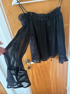 Svart offshoulder blus med volangärm - Svart offshoulder blus i transparent, lätt material med tunna axelband och långa, vida ärmar med volangdetalj vid ärmslutet. 