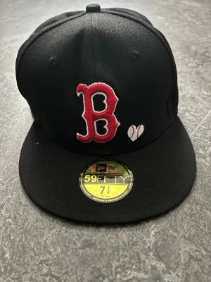 Svart Boston Red Sox keps New Era - Säljer en svart keps från New Era, modell 59FIFTY, med Boston Red Sox logga broderad i rött och vitt framtill. Kepsen har platt skärm och World Series-märke på sidan. Klassisk basebollstil och tillverkad i textil.