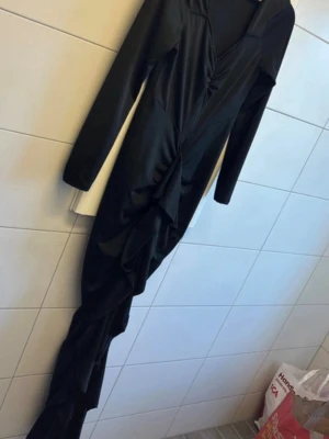 Svart långklänning med drapering - Svart långklänning med snygg drapering framtill och v-ringad hals. Klänningen har långa ärmar och en figurnära passform som framhäver siluetten. Perfekt för dig som vill ha en elegant och stilren look.Jätte fint skick. Vet ej vart den är från egen då det inte finns någon lapp men passar en M/L då den är väldigt stretchig