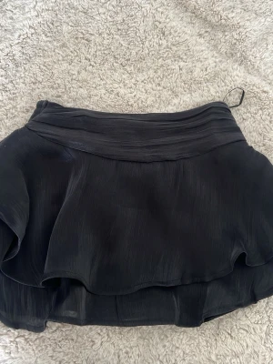 Svart volangkjol med omlottkänsla - Supersnygg svart kortkjol med dubbla volanglager och omlottkänsla. Kjolen har bred linning och ett lätt, flygigt tyg som ger en cool och trendig look. Perfekt att styla med en croppad topp eller stickad tröja.
