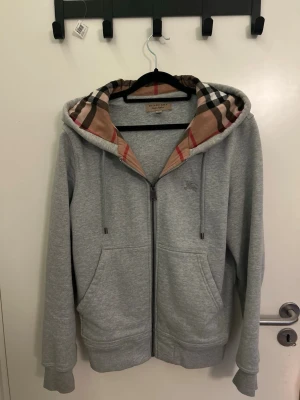 Grå hoodie från Burberry med mönstrad huva - Snygg grå zip-hoodie från Burberry med klassiskt rutmönster i beige, svart och rött på insidan av huvan. Tröjan har dragkedja, två fickor fram och ribbade muddar. Perfekt för en stilren och trendig look. Storlek S men passar även M