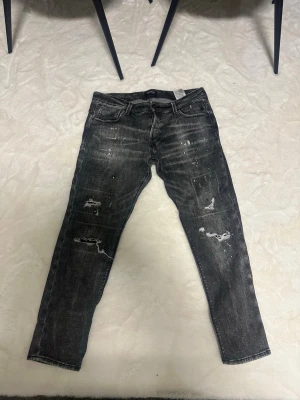 Svarta slitna jeans med distressed detaljer - Säljer ett par svarta jeans med coola slitningar och distressed detaljer på både fram- och baksida. Jeansen har fem fickor och en smal passform. Perfekta för dig som gillar en edgy streetstil. Perfekt inför sommaren bra skick på dem 2 gånger använda storlek 36 längden 32
