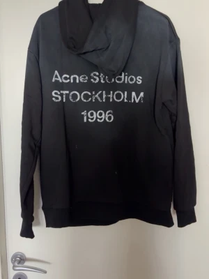 Svart hoodie Acne Studios Stockholm - Snygg svart hoodie från Acne Studios med texten 'Acne Studios STOCKHOLM 1996' på ryggen. Hoodien har en klassisk huva och raka ärmar. Perfekt för dig som gillar stilrena och trendiga plagg med streetkänsla.