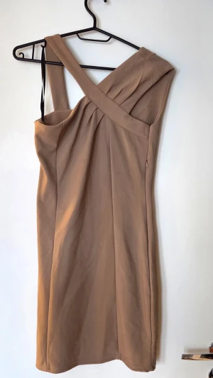 Beige one shoulder klänning med prislapp - Snygg beige kortklänning med one shoulder-design och bred axelrem. Klänningen har en figurnära passform och stilrena sömmar som ger en elegant siluett. Perfekt för dig som vill sticka ut med en modern och trendig look.