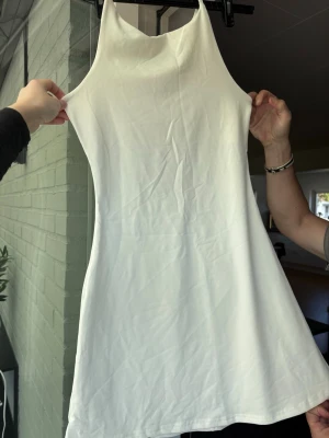 Vit ärmlös kortklänning - Snygg vit kortklänning med ärmlös design och hög halsringning. Klänningen har en enkel och stilren look, perfekt för varma dagar. Materialet känns mjukt och lätt, vilket gör den bekväm att bära. Passar dig som gillar minimalistisk stil.