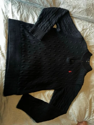 Svart kabelstickad tröja Polo Ralph Lauren - Svart kabelstickad tröja från Polo Ralph Lauren med röd broderad logga på bröstet. Tröjan har rund hals, långa ärmar och ribbstickade muddar vid ärmslut och nederkant. Perfekt för lager-på-lager och klassisk stil.