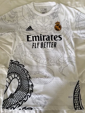 Real Madrid x Adidas draktröja - Snygg vit Real Madrid-tröja från Adidas med grått drakmönster över hela plagget. Tröjan har klubbmärke, Adidas-logga och Emirates Fly Better-tryck på bröstet. Tillverkad i lätt och ventilerande material, perfekt för fotboll eller träning.