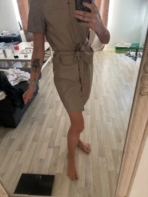 Beige skjortklänning med knytband - Snygg beige skjortklänning med korta ärmar och knytband i midjan. Klänningen har knappar framtill och en klassisk krage. Perfekt för en trendig och avslappnad look.