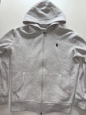 Ljusgrå hoodie från Polo Ralph Lauren - Snygg ljusgrå hoodie med dragkedja från Polo Ralph Lauren. Klassisk design med broderad logga på bröstet, två fickor framtill och mjukt material. Perfekt för en chill och stilren look.