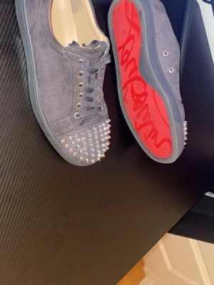 Christian Louboutin svarta sneakers med nitar - Svarta sneakers i mocka från Christian Louboutin med ikonisk röd sula och silvriga nitar på tån. Skorna har svarta skosnören, metallfärgade öljetter och låg profil. Perfekt för dig som vill sticka ut med en edgy och lyxig stil.