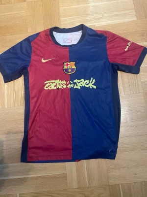 FC Barcelona x Cactus Jack matchtröja - Säljer en FC Barcelona matchtröja i rött och blått från Nike, med Cactus Jack-logga på bröstet och UNHCR-tryck på sidan. Tröjan har klubbmärke, gul Nike-logga och korta ärmar. Perfekt för dig som älskar fotboll och streetwear.
