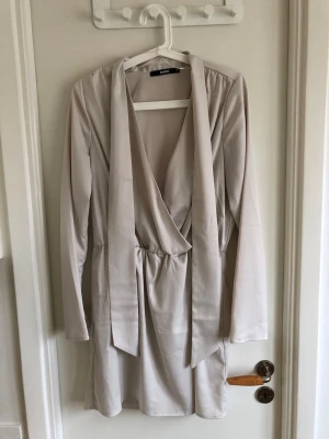Ljus omlottklänning från BikBok - Snygg ljus omlottklänning från BikBok med lång ärm och knytband i midjan. Klänningen har en djup v-ringning och är tillverkad i ett mjukt, slätt material som ger en elegant look.