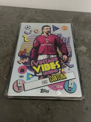 Topps Match Attax Vintage Vibes-kort - Coolt samlarkort från Topps Match Attax-serien med motiv av Eric Cantona i Manchester United. Kortet har färgglad retrodesign med texten 'Vintage Vibes Legend' och klubbens logga. Baksidan visar Manchester United och turneringsloggor. Perfekt för fotbollsfans!