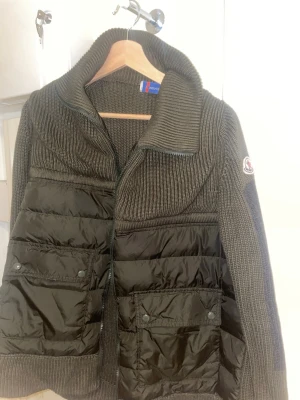 Pufferjacka i stickad design - Snygg pufferjacka från Moncler i mörkgrönt med stickade partier och vadderade paneler. Jackan har hög krage, dragkedja och två stora fickor framtill. Moncler-logga på ärmen. Perfekt för kalla vinterdagar och riktigt trendig look. Den är storlek Large men sitter mer som XL. Säljes pga gått ner i vikt och inte kan använda längre.
