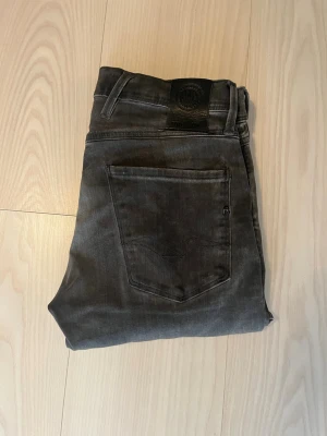 Grå slim fit jeans från Replay - Snygga grå slim fit jeans från Replay med klassisk femficksdesign och svarta detaljer. Jeansen har en smal passform och är tillverkade i ett stretchigt material som ger en bekväm känsla. Perfekta för en trendig och avslappnad stil.