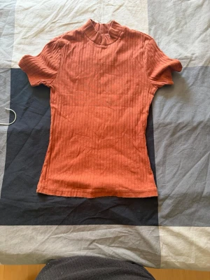Rostfärgad ribbad topp med kort ärm - En snygg rostfärgad ribbad topp med korta ärmar och liten ståkrage. Toppen har en slim passform och är tillverkad i mjuk bomull. Perfekt att styla med jeans eller kjol för en trendig look.