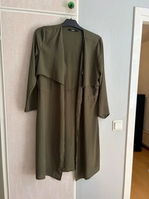 Olivgrön trenchcoat från Bik Bok - Stilren olivgrön trenchcoat från Bik Bok med lång, rak passform och öppen front. Jackan har långa ärmar och ett klassiskt fall utan knappar eller bälte. Tillverkad i ett lätt, mjukt tyg som ger en avslappnad look. Perfekt för lager-på-lager-stil.