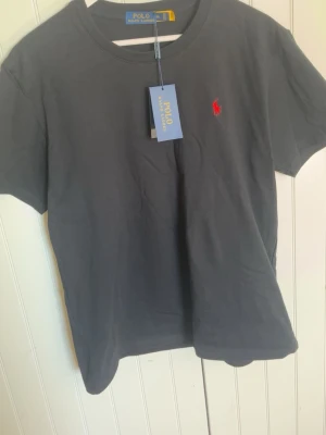 Svart t-shirt från Polo Ralph Lauren - Klassisk svart t-shirt från Polo Ralph Lauren med röd broderad logga på bröstet. T-shirten har rund halsringning och korta ärmar. Tillverkad i mjuk bomull för en skön känsla och stilren look.