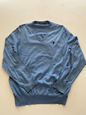 Blå V-ringad tröja Polo Ralph Lauren - Snygg blå V-ringad tröja från Polo Ralph Lauren i mjuk bomull. Tröjan har långa ärmar, ribbade muddar och den klassiska broderade loggan på bröstet. Perfekt för en stilren och avslappnad look.