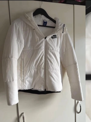 Vit pufferjacka från Nike med huva - Säljer en snygg vit pufferjacka från Nike med dragkedja framtill och huva. Jackan har en broderad Nike-logga på bröstet och justerbar dragsko i huvan. Perfekt för kalla dagar och riktigt skön att bära. Passar dig som gillar sportig och clean stil. Passar mer vår/höst