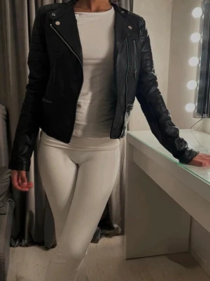 Svart skinnjacka Zara - Svart skinnjacka från Zara med silvriga dragkedjor och detaljer. Jackan har quiltade partier på axlar och ärmar samt två snedställda fickor med dragkedja. Kort, figurnära modell som ger en cool look. Liten i storleken 