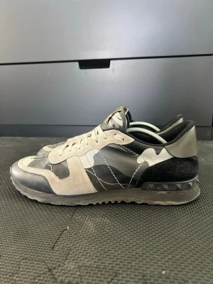 Valentino sneakers med camomönster - Snygga sneakers från Valentino i mycket fint skick. Skorna har detaljer i mocka och läder, svarta nitar på hälen och svarta samt gråa partier. Klassisk låg modell med snörning och bekväm sula. Perfekta för dig som gillar streetstyle och exklusiva märken. Priset går att diskutera vid snabb affär! Hör gärna av dig om du undrar något.