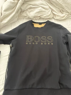 Svart sweatshirt från Hugo Boss - Snygg svart sweatshirt från Hugo Boss med guldfärgad logga framtill och dekorativa band med BOSS-tryck längs sidorna. Tröjan har rund halsringning och ribbade muddar vid ärmslut och nederkant. Perfekt för en stilren och trendig look.