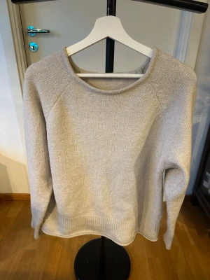 Beige stickad tröja med rund hals - En stilren beige stickad tröja med rund halsringning och långa ärmar. Tröjan har en enkel och klassisk design med ribbstickade muddar vid ärmslut och nederkant. Perfekt för lager-på-lager och passar till det mesta.