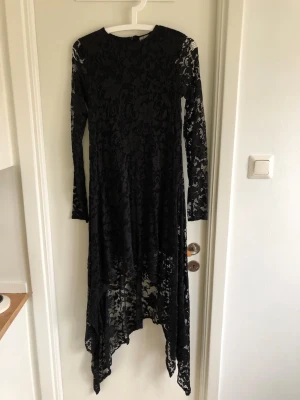 Svart spetsklänning från H&M Divided - Svart långklänning från H&M Divided i genombruten spets med lång ärm och asymmetrisk nederkant. Klänningen har rund halsringning och ett elegant blommigt spetsmönster som ger en cool och stilren look.