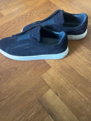Mörkblå sneakers från Axel Arigato - Snygga mörkblå sneakers från Axel Arigato i mocka med vit sula. Modellen är low top och har minimalistisk design utan snörning, med elastiska paneler för enkel påtagning. Perfekta för en clean och stilren look.