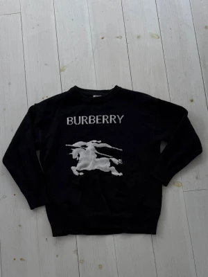 Svart stickad Burberry-tröja - Svart stickad tröja från Burberry med vit logotyp och en stor riddarillustration framtill. Tröjan har rund halsringning, långa ärmar och ribbade muddar. Perfekt för dig som gillar klassiska designer med en modern twist.