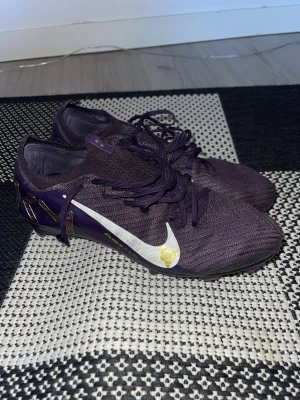 Nike Mercurial fotbollsskor lila - Säljer ett par Nike Mercurial fotbollsskor i en cool lila färg med vita och guldiga detaljer. Skorna har snörning och en lätt, stickad ovandel som ger bra passform. Yttersulan är designad för optimal grepp på planen och har en unik gul och svart design.