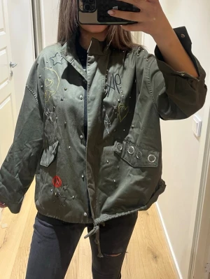 Olivgrön vindjacka med broderier - Cool olivgrön vindjacka med broderade detaljer som hjärtan, stjärnor och små nitar. Jackan har dragsko i midjan, stora fickor med metalldetaljer och tryckknappar vid ärmsluten. Oversized passform och streetig vibe, perfekt för lager-på-lager-stil.
